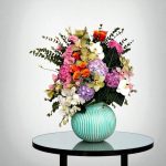 گل رومیزی فیروزه ای فلورانس با طراحی خاص خود، شامل گل‌های اورکید سفید و صورتی و رزهای هلویی، در گلدانی فیروزه‌ای مدرن قرار گرفته است. این گلدان زیبا برای هر فضایی، از دکوراسیون منزل تا محل کار، ایده‌آل است.